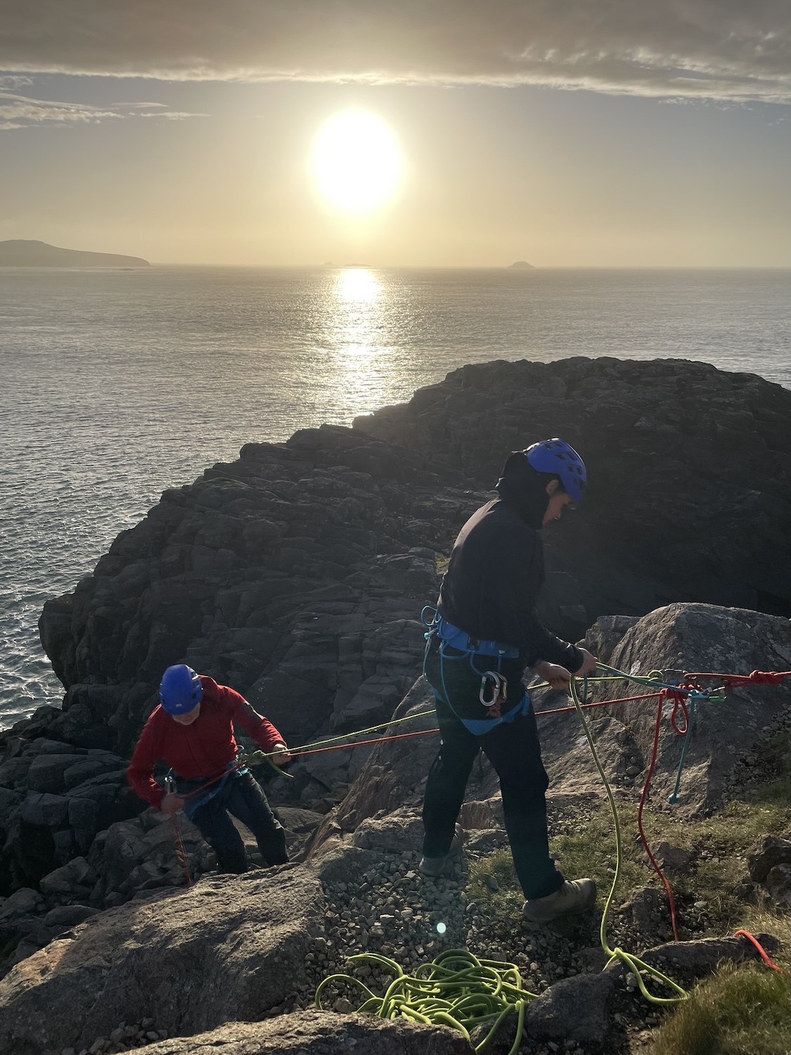 Outdoor instructor training students setting up rope systems and belays on a coastal cliff at sunset